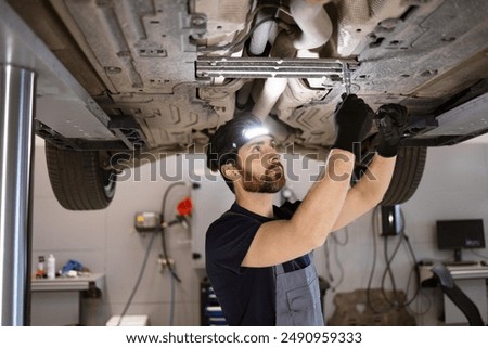 Dedicated mechanic performing maintenance on car in professional garage. Wearing headlamp and gloves, focusing on vehicle's undercarriage for repair work.