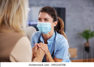Eine engagierte Krankenschwester mit Gesichtsmaske, die zu Hause mit einer älteren Frau sitzt, ihre Hände hält und sie beruhigt.