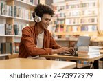 Dedicated smiling black male student engaged in online learning at college library, using laptop to study, surrounded by bookshelves
