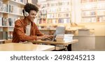 Dedicated smiling black male student engaged in online learning at college library, using laptop to study, surrounded by bookshelves