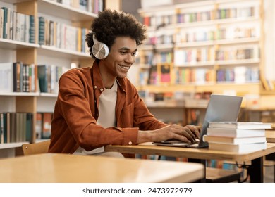 Dedicated smiling black male student engaged in online learning at college library, using laptop to study, surrounded by bookshelves - Powered by Shutterstock