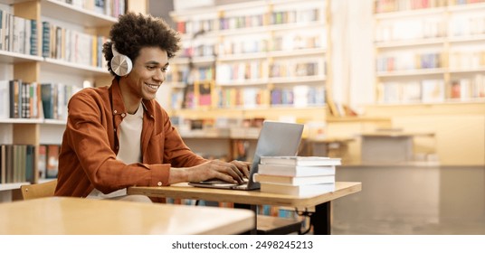 Dedicated smiling black male student engaged in online learning at college library, using laptop to study, surrounded by bookshelves - Powered by Shutterstock