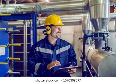 Dedicated smiling factory worker standing next to boiler and holding tablet. Worker is dressed in protective uniform, having hardhat and antiphons.