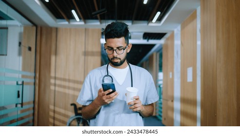 Dedicated uniformed and professional male nurse using smartphone and internet and walking in the corridors of hospital during break. Successful young worker drinking coffee. - Powered by Shutterstock