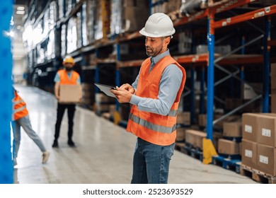 A dedicated warehouse worker stands in a bright stockroom, using a digital tablet for task analysis. His safety helmet and vest highlight his commitment to efficient inventory management. - Powered by Shutterstock