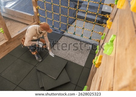 A dedicated worker is focused on laying down safety flooring in a children's play area at a recreational facility. Brightly colored climbing holds are visible on the wooden wall, while safety mats