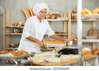 Dedicated young female baker in white chef uniform portioning and weighting dough on floured work surface in inviting artisan bakery against backdrop of baked goods arranged on wooden shelv - Powered by Shutterstock