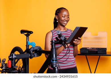 Dedicated young woman repairing a broken bicycle with tools and technology, against an isolated yellow background. Female african american cyclist thoroughly inspecting bicycle with digital tablet. - Powered by Shutterstock