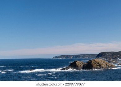 The deep blue Atlantic Ocean with waves crashing on a rocky island. The sea's coastline has rugged high cliffs with hills covered in hills. The sky is blue with a pink hue in the low lying clouds.  - Powered by Shutterstock