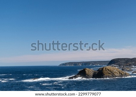 The deep blue Atlantic Ocean with waves crashing on a rocky island. The sea's coastline has rugged high cliffs with hills covered in hills. The sky is blue with a pink hue in the low lying clouds. 