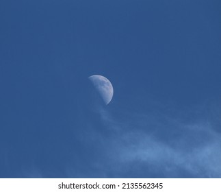The deep blue evening sky with the moon, bright gray with dark spots and craters with small wisps of clouds flowing around it.