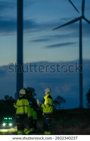 In deep blue twilight, two technicians dwarfed by the machines stand small beneath the huge dark towers at the foot of towering wind turbines. Clean energy production, field engineering and.