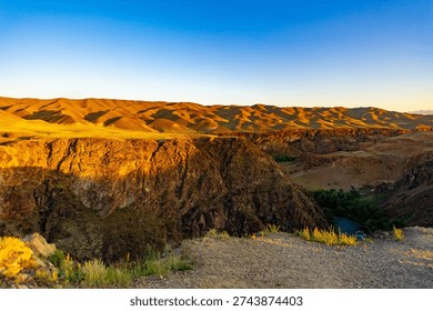 A deep canyon with a thin river at the bottom is flanked by massive rock faces and rolling golden hills under a clear sky.