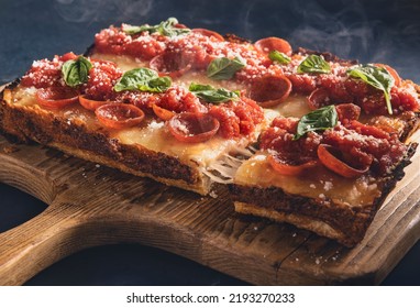 Deep Dish Rectangular Pepperoni Pizza With Basil And One Slice Being Taken Out Showing A Cheese Pull.  Pizza Sits On A Rustic Paddle Style Cutting Board