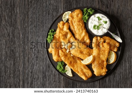 deep fried crispy beer batter fish fillet on black plate with tartar sauce and lemon slices on dark wooden table, horizontal view from above, flat lay, free space