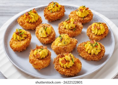 Deep fried deviled eggs with crispy coating of panko and parmesan, sprinkled with fresh chives and crumbled bacon on white plate on wooden table, close-up