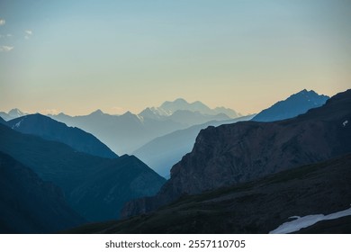 Deep gorge between layered silhouettes of sharp rocky ridge spurs under huge snow-capped mountain peak in golden blue gradient sky. Sheer crags of mount valley under giant ice top of gold sunset color - Powered by Shutterstock