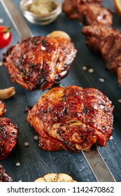 Deep Grilled Hot And Spicy Bbq Chicken Thighs On A Skewer Close Up. Macro Photo Of Appetizing Barbecue Poultry Legs With Spices, Tomatoes, Garlic, Salt On Black Blur Background