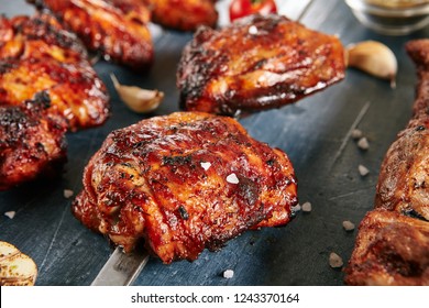 Deep Grilled Hot And Spicy Bbq Chicken Thighs On A Skewer Close Up. Macro Photo Of Appetizing Barbecue Poultry Legs With Spices, Tomatoes, Garlic, Salt On Black Blur Background