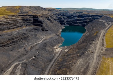 Deep opencast mine pit filled with turquoise water at bottom, terraced coal seam slopes and haul dirt roads surrounding flooded void on clear bright day