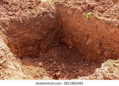 Deep rectangular foundation trench dug by excavator in raw clayey soil, showing textured earthen walls and loose dirt at the bottom on an active construction site development zone