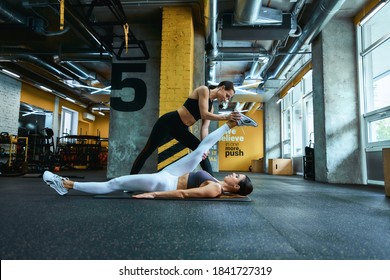 Deep stretch. Side view of a young beautiful fitness woman lying on yoga mat at gym and doing stretching exercises with assistance of her female personal trainer - Powered by Shutterstock