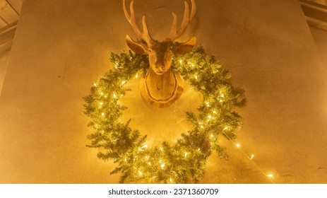 Deer head with glowing Christmas wreath hangs on wall. Circle garland made of fir twigs and fairy lights in living room. Traditional Xmas holiday ornament in semi-dark room. New year celebration - Powered by Shutterstock