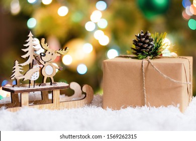 Deer In A Sleigh And A Box With A Gift Close-up On A Bokeh Background. Copy Space, Front View. New Year And Christmas Background For Greetings, Banner, Postcard, Advertisement With Place For Text.