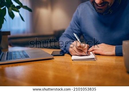 Define goals, write tasks, do a checklist. A happy young businessman is sitting at home, working remotely, and writing tasks in his spiral block. There is a laptop on a table.