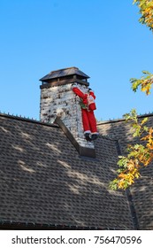 Deflated Blow Up Santa Tied To Chimney Looking Limp And Like He Is Trying To Hang On - Funny - With Fall Leaves And Light Bulbs On Roof Line - Early Decorating For Christmas