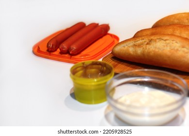 Defocus Ingredients For Making Homemade Hot Dogs. Sausages In Orange Plate, Fresh Baked Buns, Mustard And Sauce, Served On White Background.  Hotdog With Different Ingredients. Out Of Focus.