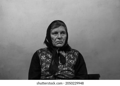 Defocus Portrait Of 60s Russian Grandmother Senior Old Woman Seating Indoors. Old Women Shawl. Sad Person. White Dirty Background. Black And White. Out Of Focus.