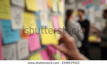 Defocused abstract background of a creative workspace. A hand places a yellow sticky note on a wall filled with colorful notes, sketches, and doodles.
