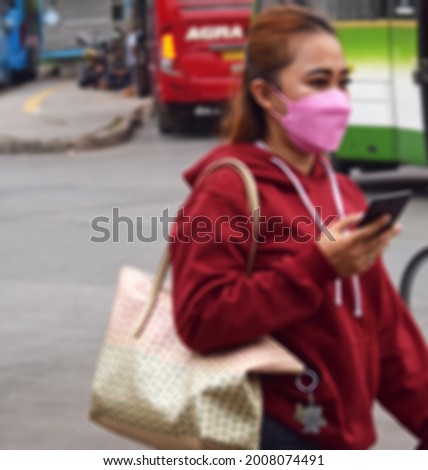 Defocused abstract background of Indonesian girl wearing pink fac mask against disease virus SARS-CoV-2. Girl with face mask walks holding mobile phone during Pandemic Corona virus Disease 2021