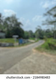 Defocused Abstract Background Of A Street Scene With A River In One Corner Decorated With Trees, People's Houses, And A Cloudy Blue Sky