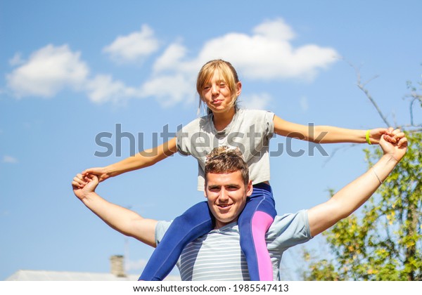 Defocused brother riding sister on back. Portrait\
of happy girl on man shoulders, piggyback. Girl fly, raise hands.\
Family playing outside. Blue green nature background. Summer mood.\
Out of focus.