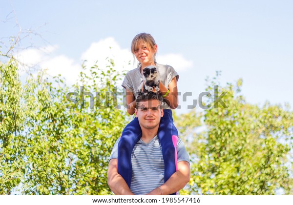 Defocused brother riding sister on back. Portrait\
of happy girl on man shoulders, piggyback. Girl holding cat,\
kitten. Family playing outdoor. Pet, animal. Green background. Copy\
space. Out of focus.