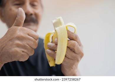 Defocused happy senior man wwith thumb up and isolated on a white background holds a peeled banana in his hand ready to be eaten - Powered by Shutterstock