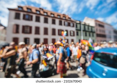 Defocused Large Crowd Of Participants At Annual Gay Pride With Flags And Other Memorabilia Walking City Center With Tall Houses In Background Blue Sky