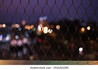 Defocused Night Light City Scape Through Metal Grid, The Sky Are Purple, Blue, And Pink With Colourful Bokeh Are Yellow Green Red And White.