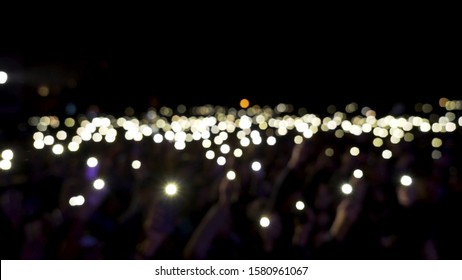 Defocused Spectators In Hall With Lights Waving In The Dark. Action. Audience Holding Phones With Shining Flashlights During The Concert, Romantic Atmosphere.