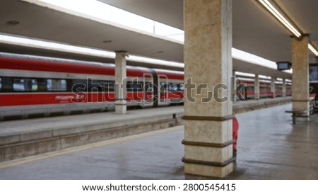 Defocused train platform scene with blurred red carriage, concrete pillars and empty tracks in soft bokeh; background copyspace backplate calm.