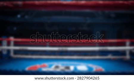 A defocused view of an empty boxing ring featuring red ropes and a blue mat under bright arena lights.