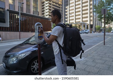 Defocused Of Young Asian Traveller Carrying A Black Bags Standing Paying Carparking By Inserting Coin Into Parking Metre Machine Perth City CBD Street In The Afternoon Light  �