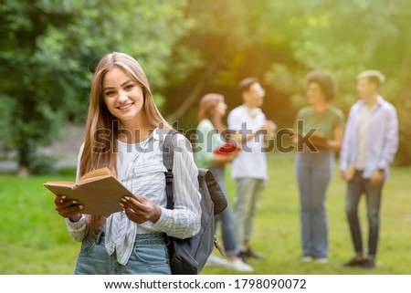 Degree Programs Abroad. Portrait Of Smiling Student Girl With Book Posing Outdoor At Campus With Her Classmates On Background, Selective Focus