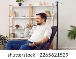 Dehydrated young man receiving intravenous vitamin therapy in hospital room. Male patient sitting in armchair attached to vitamin IV infusion drip in wellness center or beauty salon