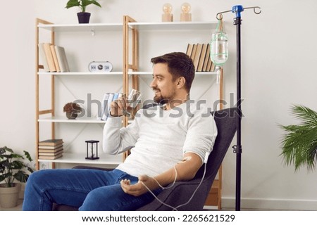Dehydrated young man receiving intravenous vitamin therapy in hospital room. Male patient sitting in armchair attached to vitamin IV infusion drip in wellness center or beauty salon