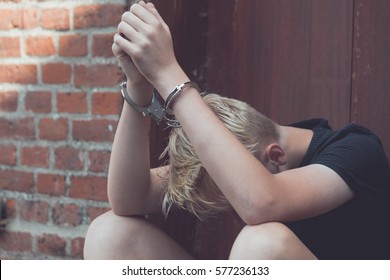 Dejected teenage boy held captive in handcuffs sitting on the ground against an exterior wooden door with his head down between his legs after being busted and arrested - Shutterstock ID 577236133