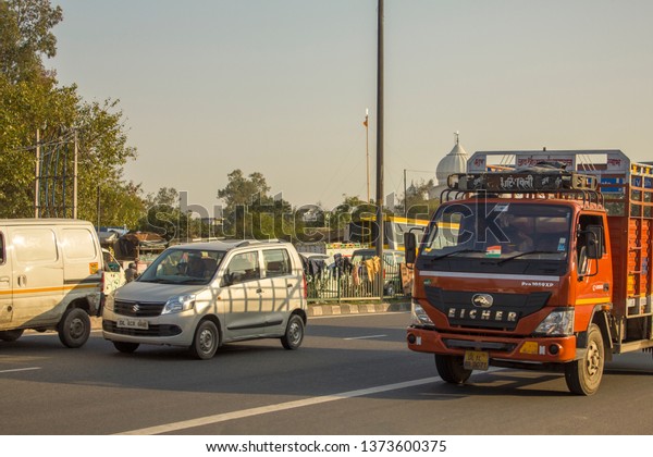 Delhi/ India - 09.02.2019: Indian red\
truck and gray passenger car in city traffic en against the\
background of green trees and the dome of the white\
temple
