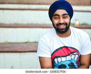 Delhi, India 10/01/2014: Close Up Of An Indian College Boy Student In Casual Wear And Blue Turban Smiling While Sitting Outdoors On A Sunny Morning In The University Campus.